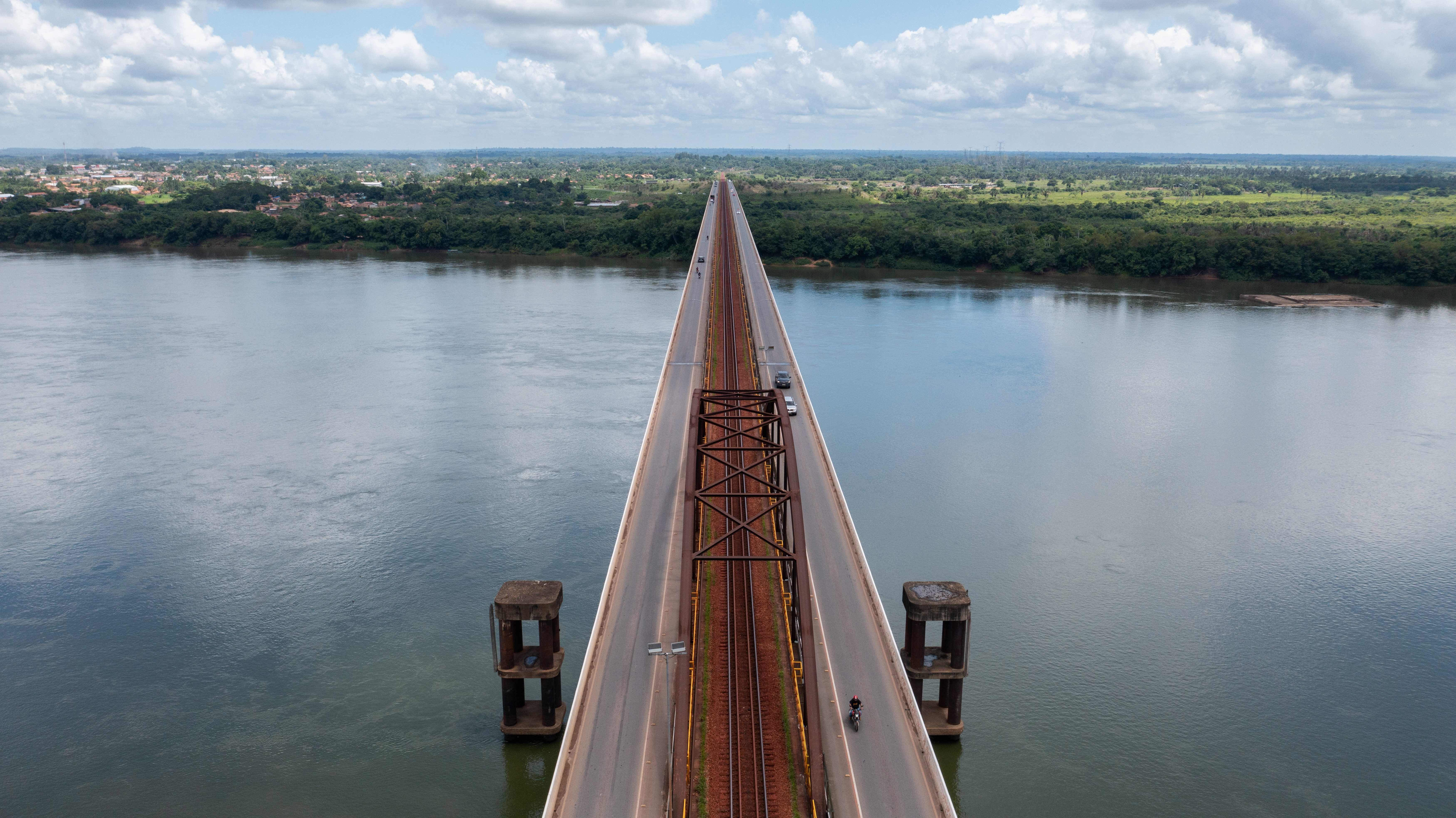 Foto da ponte vista de cima em cima de um rio e uma vegetação ao redor do rio. A ponte possui uma pista para cada sentido e no meio um trilho para trem. 