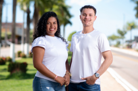 Uma mulher e um homem sorriem para foto. Ambos usam camisetas brancas e estão ao ar livre.