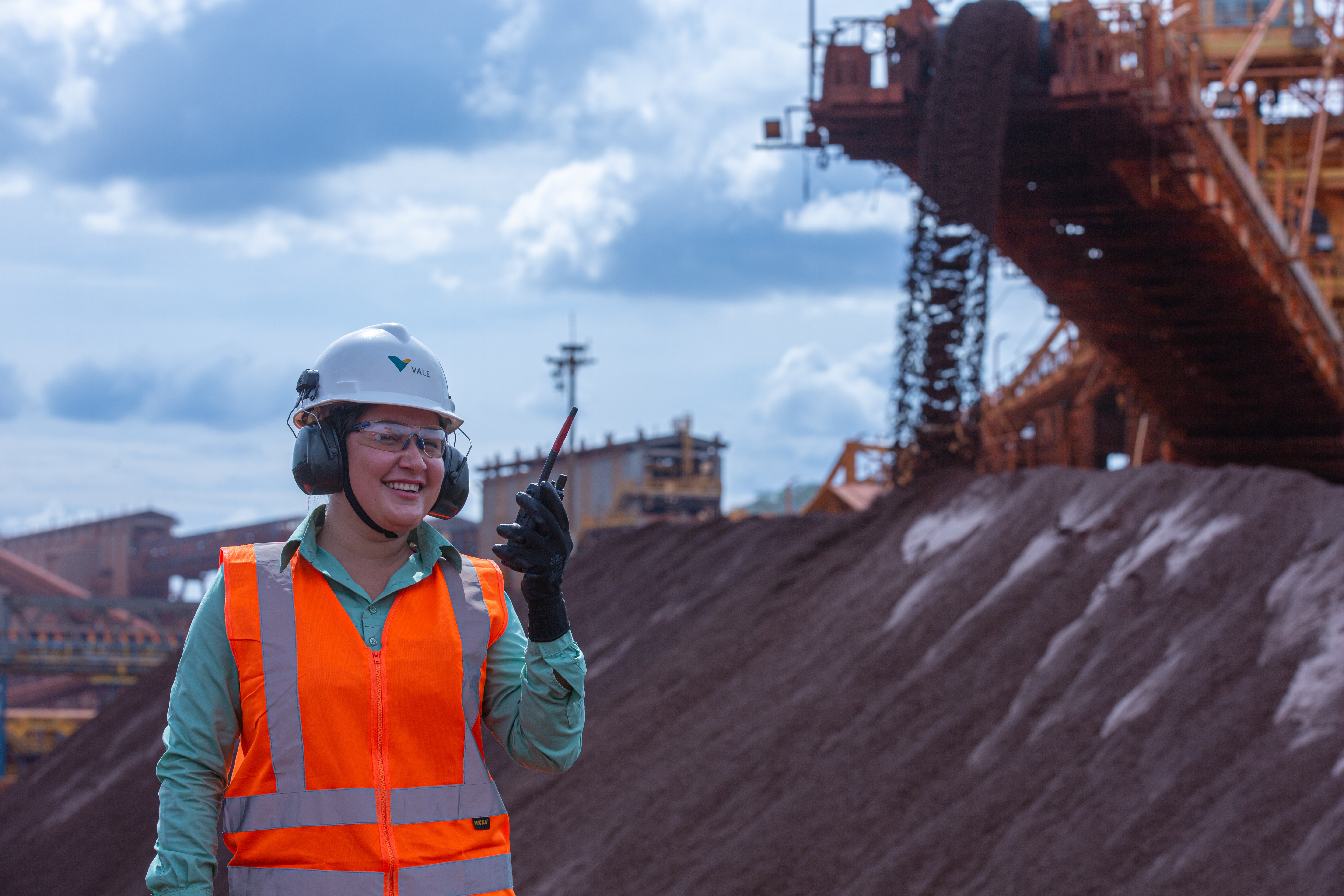 Person wearing safety gear — white helmet, earmuffs, and orange reflective vest — holding a walkie-talkie in front of a large pile of material, possibly soil or ore. In the background, there is industrial machinery and a conveyor belt system. The sky is partly cloudy.