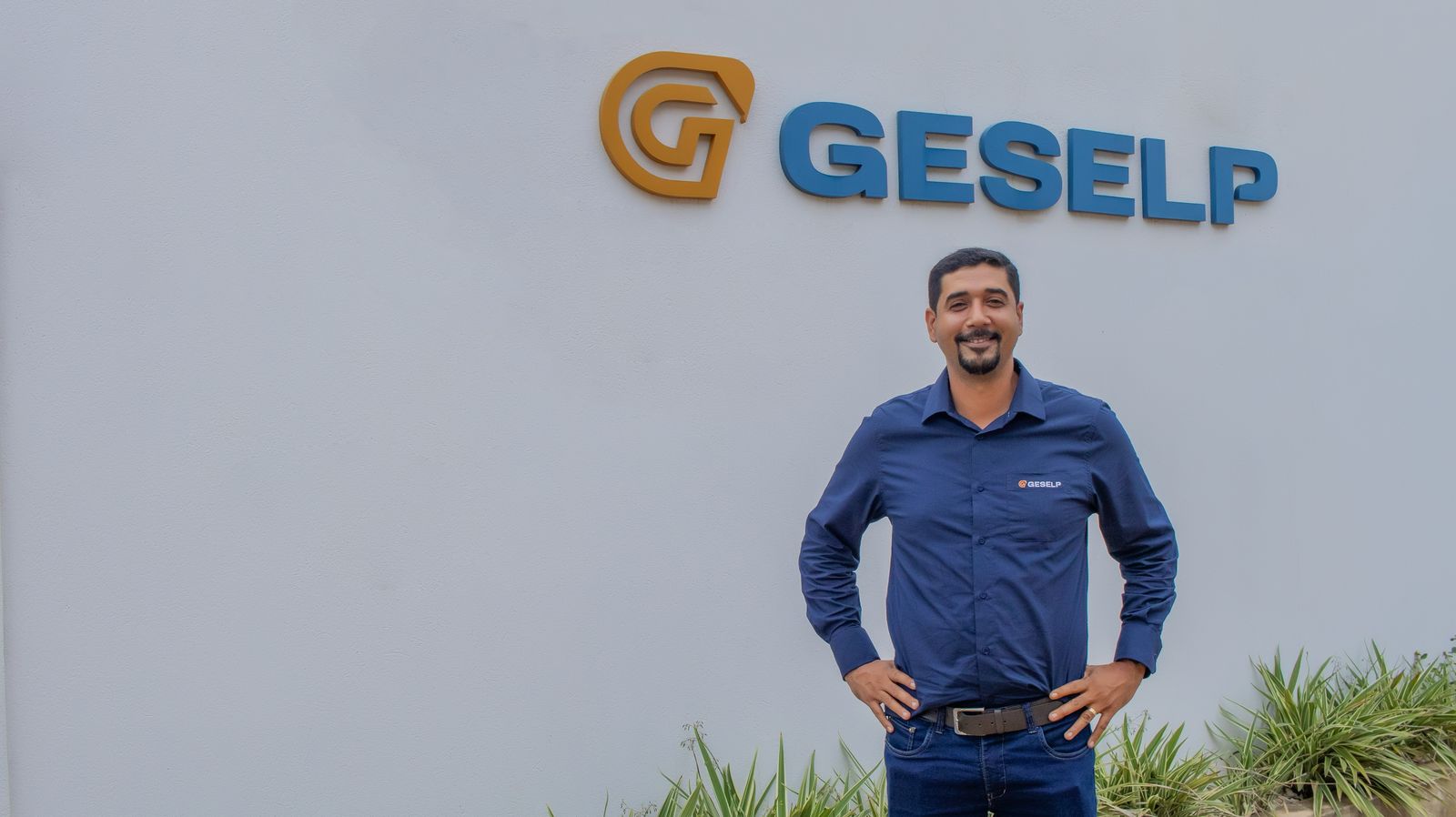 Person standing in front of a wall with the "GESELP" logo and name. They are wearing a blue shirt with the same logo on the left side of the chest. There are some plants at the bottom of the wall.