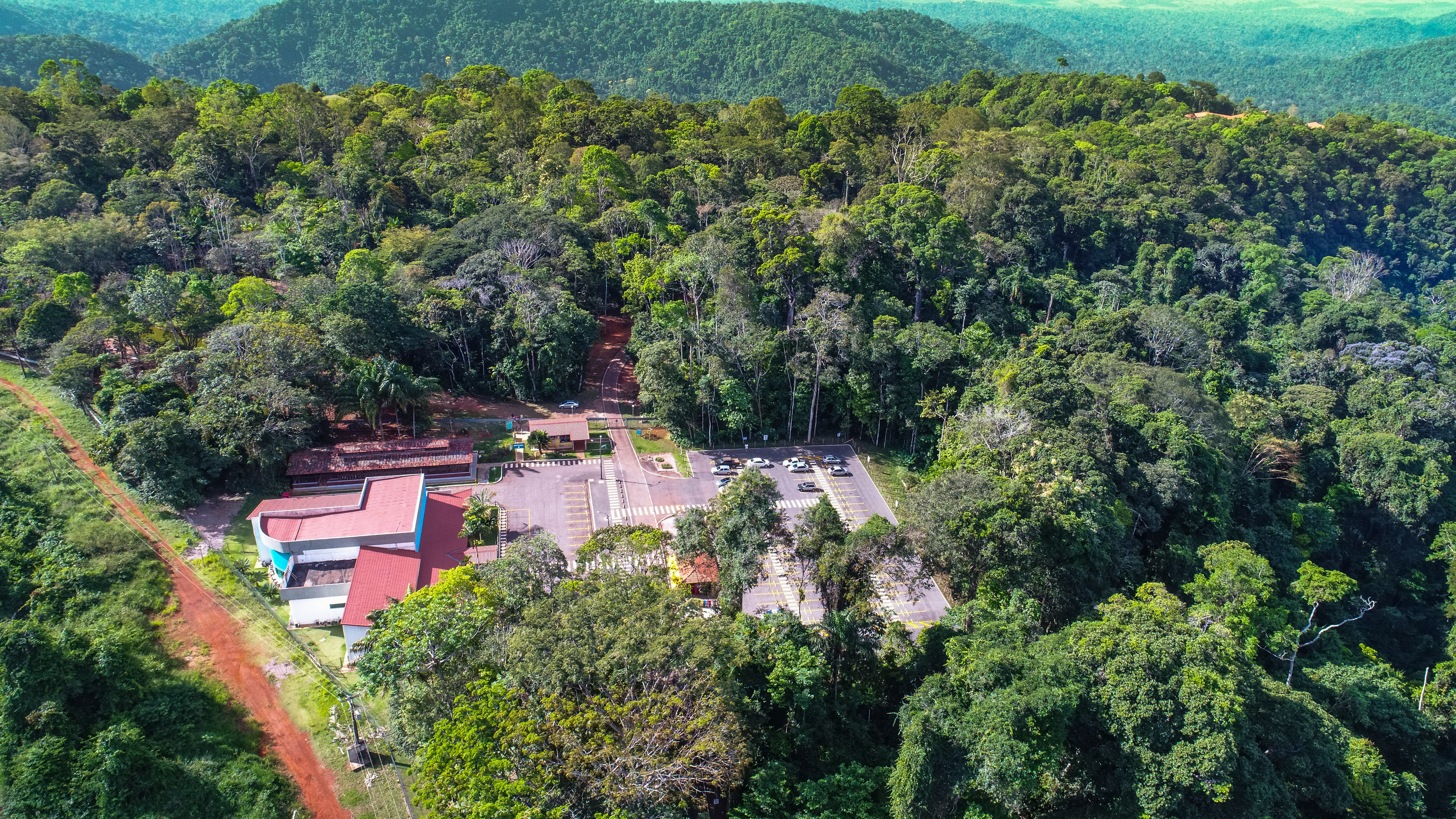 Aerial view of a building complex surrounded by dense forest. It includes structures with red and white roofs, parking lots with cars, and pathways connecting the buildings. In the background, a vast green forest.