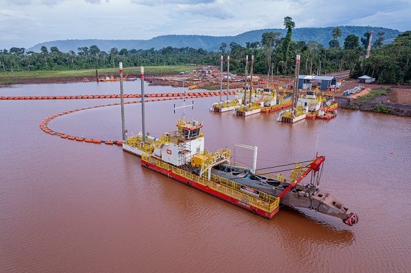 Image of a dredging operation on a river or lake. Several dredging vessels are anchored in the water, with one prominently in the foreground. The vessels are equipped with large machinery for removing sediment from the waterbed. In the background, there is a forested area and construction activity on the shore, including visible equipment and structures. Orange floating barriers are in the water, likely used to contain sediment.