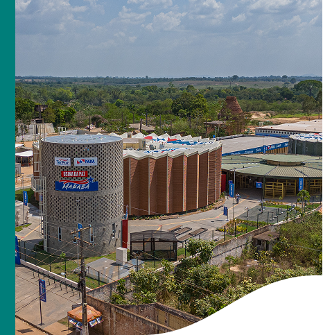 Aerial image of a building complex with a cylindrical structure in the foreground. The building displays signage reading "USINA DA PAZ MARABÁ" along with logos for the Government of Pará and the Municipality of Marabá. The area is surrounded by greenery and other buildings, with a clear sky above.