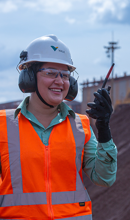 Person wearing a white hard hat with the “Vale” logo, safety glasses, ear protection, an orange high-visibility vest over a light blue shirt, and black gloves. Holding a walkie-talkie. In the background, industrial structures and an overcast sky.