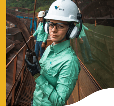 Foto de uma mulher com uniforme Vale, camisa de botões verde, colete laranja com iluminação fluorescente, capacete, máscara, óculos de proteção, luva e bota. Ao fundo tem uma roda de caminhão enorme, o dobro da altura da mulher.