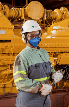 A woman in an operation area with machines in the background. She is wearing a Vale uniform, a light green and gray overalls with fluorescent markings, goggles, gloves, mask, helmet, and ear protection.