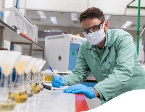 a man is in a laboratory looking at containers that are filled with a golden liquid and making notes. He is wearing a light green lab coat, gloves, face mask, and goggles.
