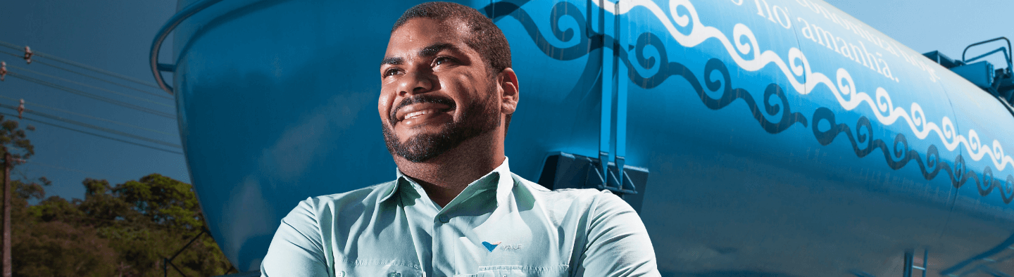 A black man smiling and looking forward. He is wearing a light green shirt with Vale logo. and behind him, there is a blue and white metal structure.