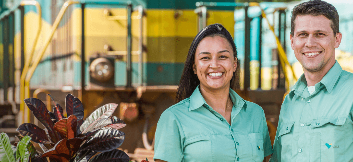 Foto tirada da cintura para cima de dois funcionários da Vale – um homem e uma mulher – sorrindo em um espaço de operações. Os dois usam camisas verdes em um tom claro.