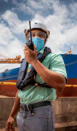 A man holds a communication radio in his hands. He is in an operational outdoor space. He is wearing a light green T-shirt, helmet, goggles, and face shield.