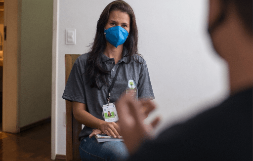 Foto de uma mulher com cabelos lisos, longos e escuros sentada em uma cadeira, olhando para frente. Ela usa máscara facial e crachá.