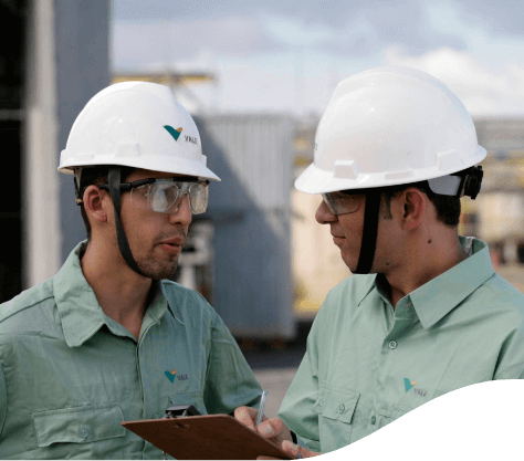 Two men talking in an operation area. One of them is holding a clipboard. Both are wearing light green Vale button-up shirts, goggles and helmets.