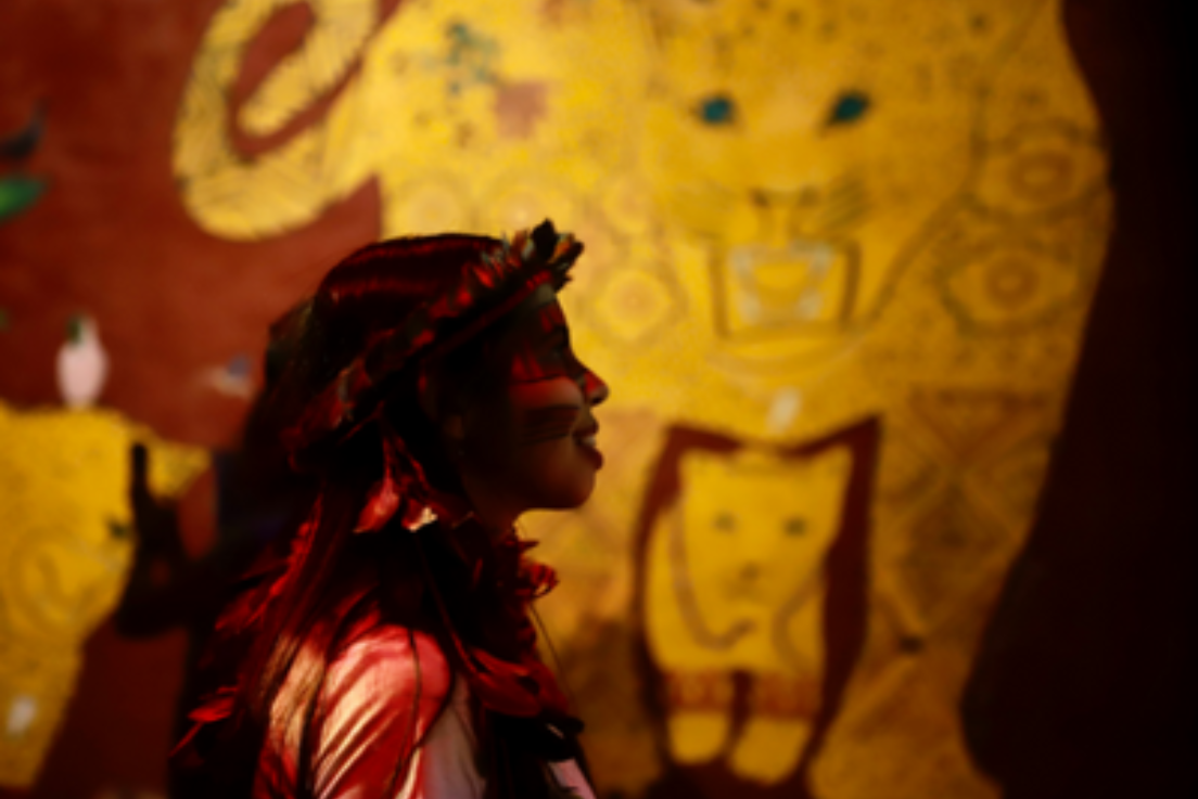 Mulher indígena aparece em primeiro plano e ao fundo está a pintura de animais na parede.