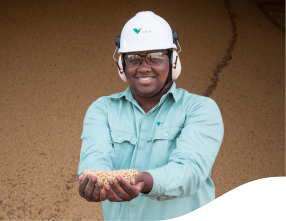 Vale employee holding grains in his hands in an operation full of grain. He is smiling and wearing a long-sleeved light green shirt, goggles, ear muffs and a white helmet with Vale logo.