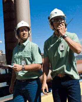 Two men side by side in an operation space. The two wear jeans, green shirts, goggles, ear muffs and white helmets with Vale logo.   One of them holds a radio communicator in one hand.