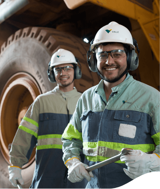 Two men smiling in an operation area with a truck wheel in the background. One of them is holding a tool. They are wearing light green and gray Vale overalls with fluorescent markings, gloves, goggles, white helmets, and ear protection.