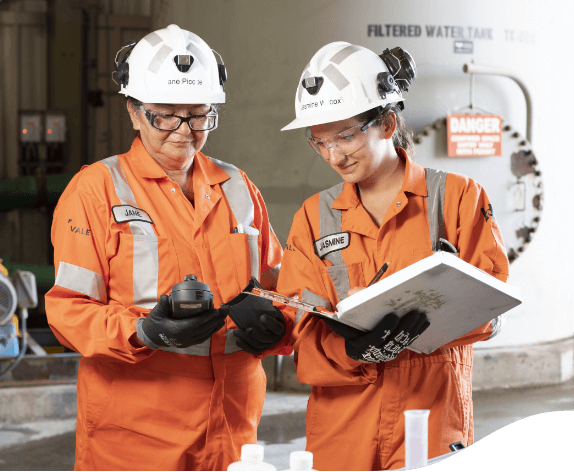 Photo of two women, one is holding a small piece of equipment and the other a notebook with a pen. The two are wearing orange overalls, gloves, white helmets, and goggles.