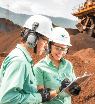 Two Vale employees, a man and a woman, analyze a clipboard. They are in the middle of an environment with earth and are wearing helmets, gloves, and goggles.