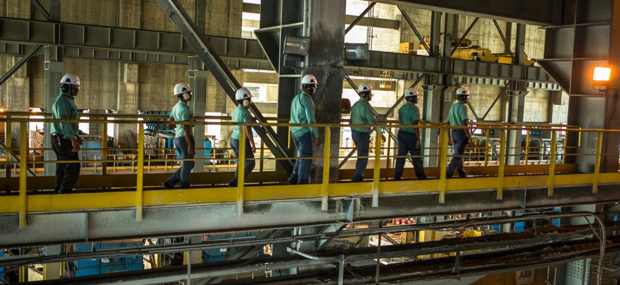 Vários empregados da Vale caminhando lado a lado em um espaço operacional. Todos usam uniformes verdes, crachá, óculos de proteção, protetores de ouvido e capacete branco com logotipo da empresa.