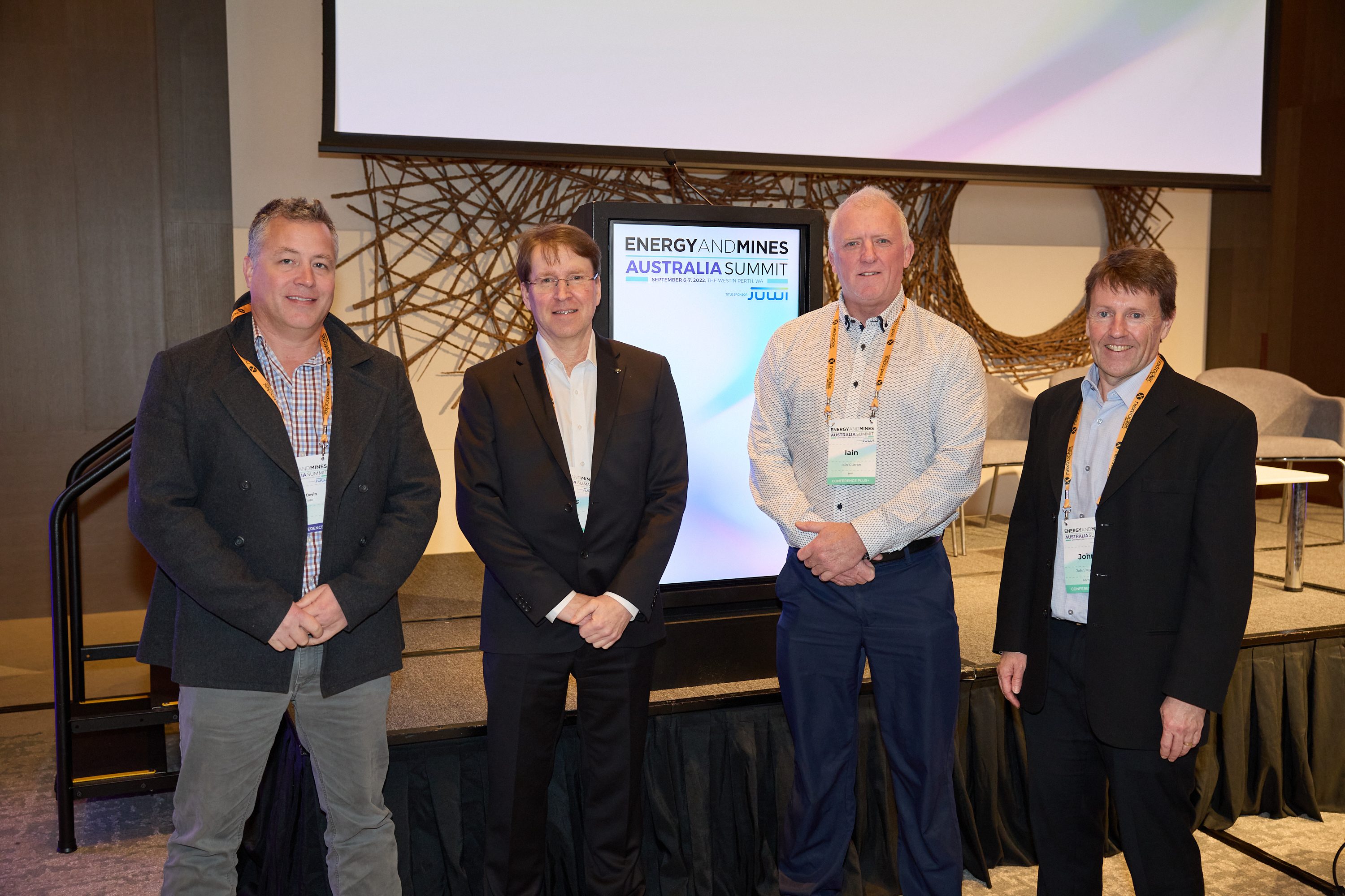 Four men stand side by side. They wear formal clothes and are standing in front of a stage, and behind them is an electronic sign that reads: “Energy and mines. Australia Summit”. 