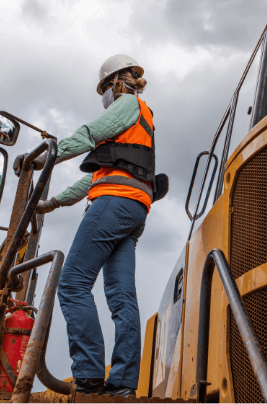 A woman is standing on top of the structure of a large vehicle in an operational area. She is wearing jeans, a light green shirt, an orange vest with gray details, hair tied back, a face mask and a white helmet.