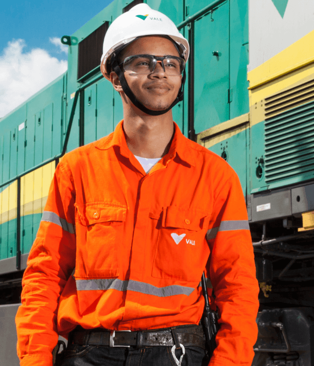 Homem parado ao lado de um trem verde, amarelo e cinza. Ele usa uniforme laranja com logotipo da Vale, óculos de proteção e capacete branco também com logotipo da empresa.