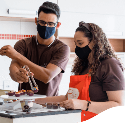 A man and a woman, both wearing brown T-shirts and protective masks, are in a kitchen making brigadeiro.