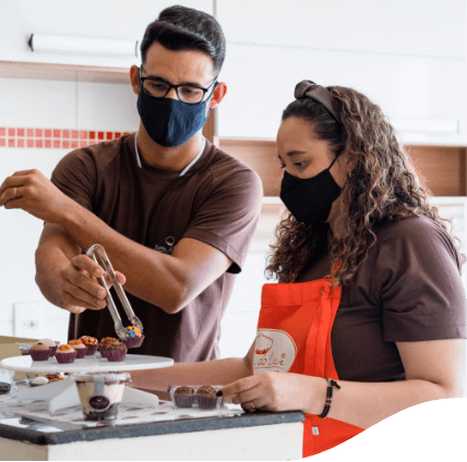 Um homem e uma mulher, ambos de camiseta marrom e máscara de proteção, estão em uma cozinha manuseando brigadeiros.