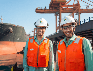 Dois homens em uma área de operação próximo ao mar estão sorrindo. Eles usam camisa em um tom de verde claro, colete laranja, capacete branco e óculos de proteção.