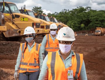 Foto da altura do peito até a cabeça de três empregados da Vale, dois homens e uma mulher. Os três estão com uniforme verde da Vale, colete laranja e cinza, óculos, capacete e máscara.