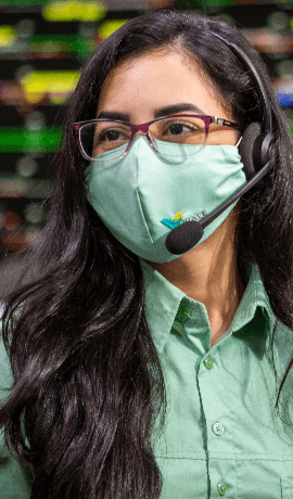 Photo of a woman with long hair in an office. She is wearing a mask, glasses, headphones with a microphone and a green button-down shirt.