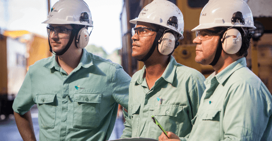 Foto de três homens observando algum ponto e uma máquina de ferro grande ao fundo. Os três estão usando camisa de botões verde clara com logo da Vale, proteção nos ouvidos, capacete e óculos de proteção. Um deles está segurando uma caneta e um papel na mão