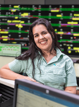 Foto de uma mulher em um escritório e vários monitores ao fundo. Ela está apoiada em um balcão e está usando camisa verde de botões com logo da Vale, cabelo comprido solt liso e está sorrindo.