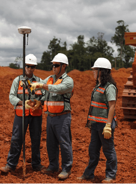 Foto de dois homens e uma mulher conversando e observando um equipamento que se assemelha a uma barra de ferro em uma área de operação. Eles estão usando camisa verde de botões da Vale, colete laranja com listras fluorescentes, calça jeans, luvas, bota, óculos de proteção e capacete.