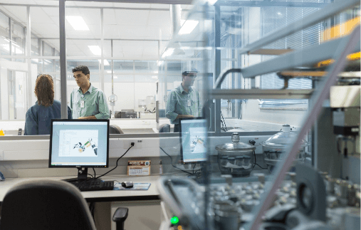 Image of a laboratory. You can see a computer with its screen on and two people talking in the background.