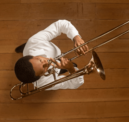 Foto tirada de cima de um músico tocando trompete. Ele usa uma camisa branca e o piso é de madeira.