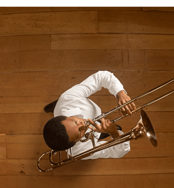 Foto tirada de cima de um músico tocando trompete. Ele usa uma camisa branca e o piso é de madeira.