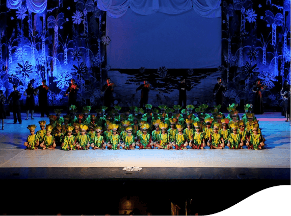 Apresentação de teatro em um palco escuro com luzes azuis. Há diversas crianças sentadas no palco, todas vestidas de verde e amarelo.