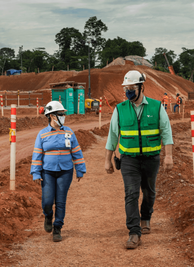 Foto de um homem e uma mulher conversando e andando em uma área de terra na operação. A mulher está usando uma camisa azul com linhas fluorescentes, calça jeans, bota máscara, óculos de proteção e capacete. Já o homem está usando camisa verde de botões da Vale, um colete verde com linhas fluorescentes, calça jeans, bota, capacete, óculos de proteção, máscara e está com um rádio preso na cintura.