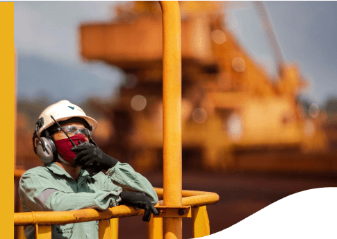 Photo of a woman in an operation area leaning against iron grid, holding and talking on a radio. She is wearing a uniform, a light green Vale shirt, gloves, ear protection, a mask, a helmet with Vale logo and goggles.