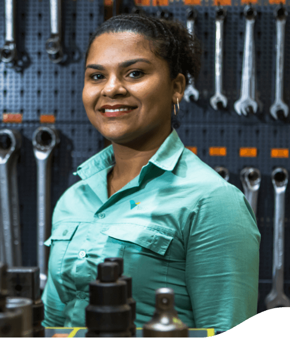 Mulher negra sorrindo em um espaço repleto de ferramentas de manutenção. Ela usa os cabelos presos e uma camisa verde clara com logotipo da Vale.