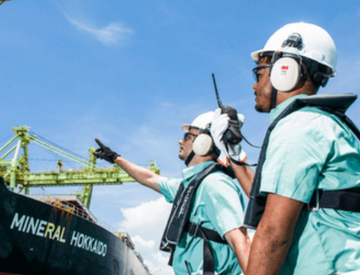 Dois empregados da Vale de frente para um navio em uma área operacional. Os dois usam camisa verde, equipamentos de proteção presos ao corpo, óculos de proteção, protetores de ouvido e capacete branco.