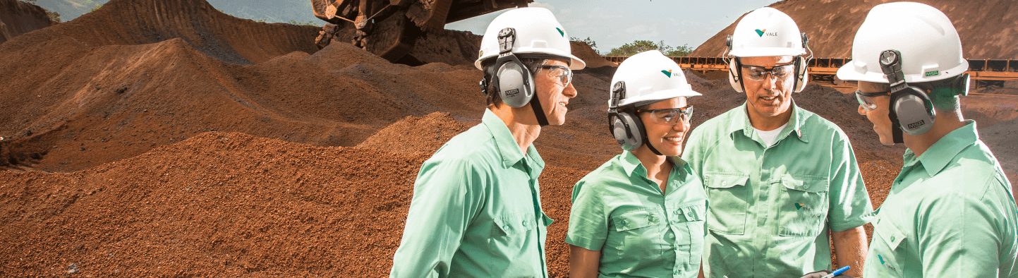 Photo of three men and a woman talking in an operation area with mountains of dirt and a machine in the background. The four are wearing uniforms, green button-down shirts, white helmets with Vale logo, goggles and ear protection.