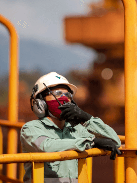 oto de uma mulher em uma área de operação apoiada em uma grade de ferro, segurando e falando em um rádio. Ela está usando uniforme, camisa verde clara da Vale, luvas, proteção nos ouvidos, máscara, capacete com logo da Vale e óculos de proteção