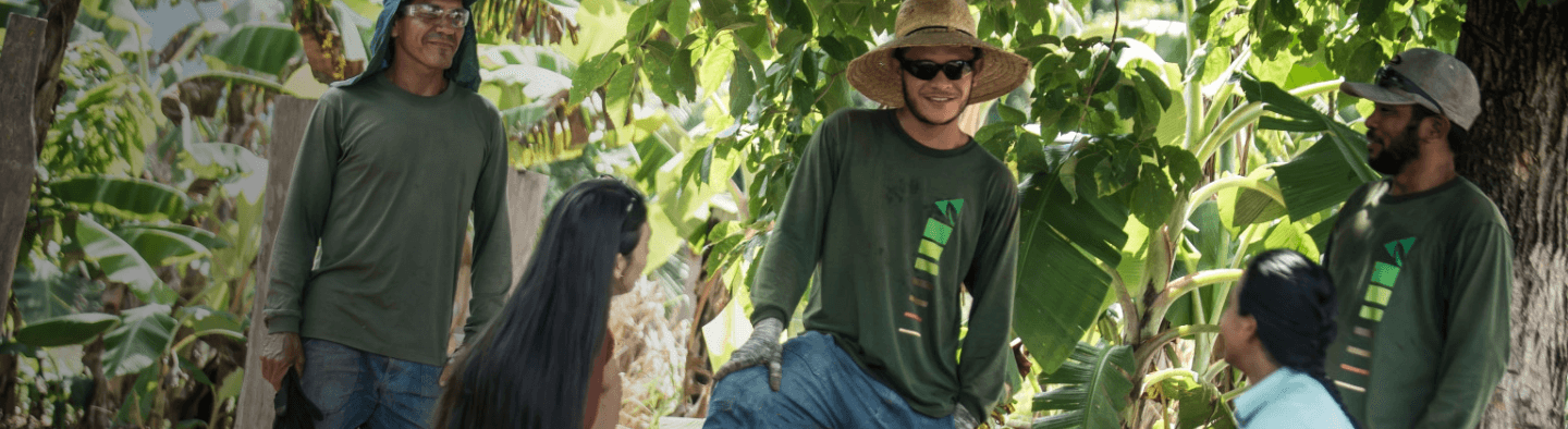 In a vegetation area, three men and two women are talking. The men are wearing dark green long-sleeved shirts, sun protection on their heads - such as a hat and cap - and are wearing  glasses and gloves.