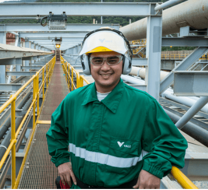 Um homem sorrindo em uma passarela de uma área de operação. Ele usa camisa verde escura, capacete branco, óculos de proteção e proteção nos ouvidos.