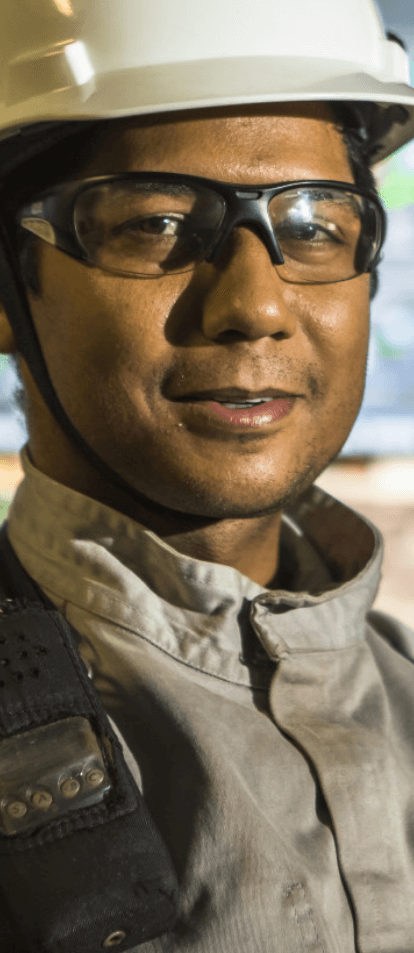 Photo from shoulder height upwards of a Vale employee. He is wearing goggles and a white helmet with the company logo. In the background are several computer screens.