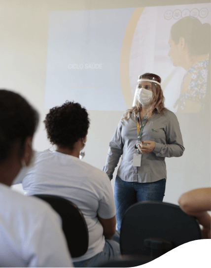 A woman in a gray shirt, jeans, a badge and two face masks making a presentation supported by a big screen, which is behind her. In front of her, there are a few people sitting watching what is being shown on the big screen.