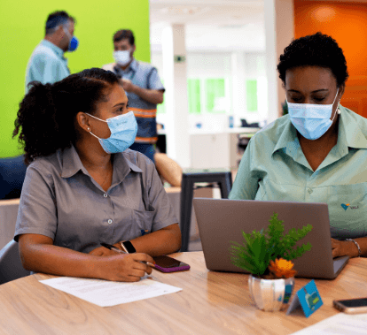 Foto de duas mulheres negras sentadas lado a lado em uma mesa. Uma delas está digitando em um notebook e as duas usam camisas de uniforme e máscaras de proteção facial.