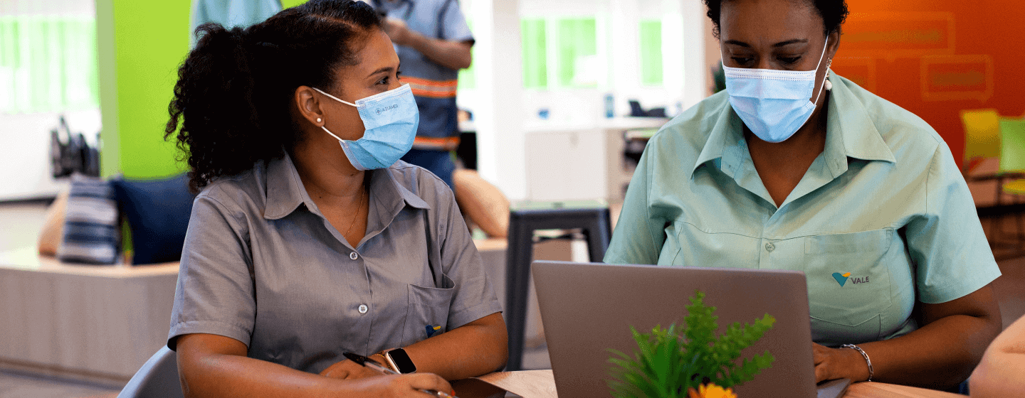 Foto de duas mulheres negras sentadas lado a lado em uma mesa. Uma delas está digitando em um notebook e as duas usam camisas de uniforme e máscaras de proteção facial.